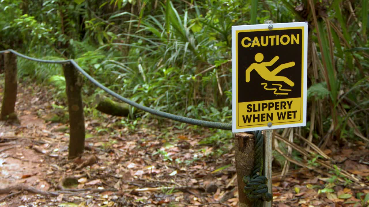 A yellow caution sign warns hikers of slippery conditions on a lush tropical forest path