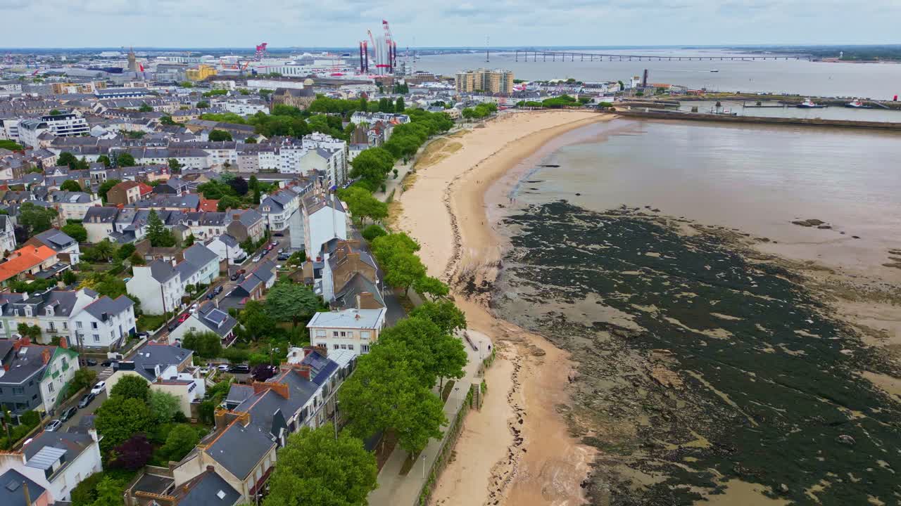 Petit Traict beach in Saint-Nazaire, cityscape, port, and Saint-Nazaire Bridge over Loire estuary in background, France. Aerial forward