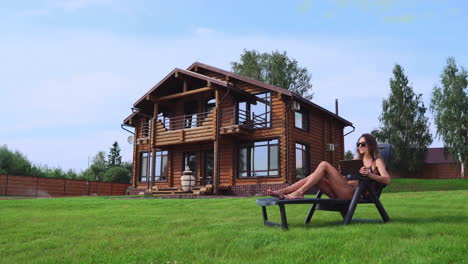 a beautiful brunette in a swimsuit lying on a sun lounger near her chic mansion with large windows smiles while working at home remotely via the internet in the business sphere. the concept of country life