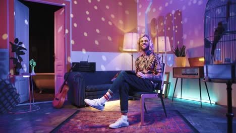 young stylish caucasian man with long hair in headphones sitting on a chair and using tablet in a retro party at home