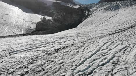 Aerial-flyover-over-a-hiker-and-mountaineer-walking-across-the-ice-and-jumping-over-the-crevasses-of-the-Allalin-glacier-near-Saas-Fee-in-Valais,-Switzerland-on-a-sunny-day-in-the-Swiss-Alps