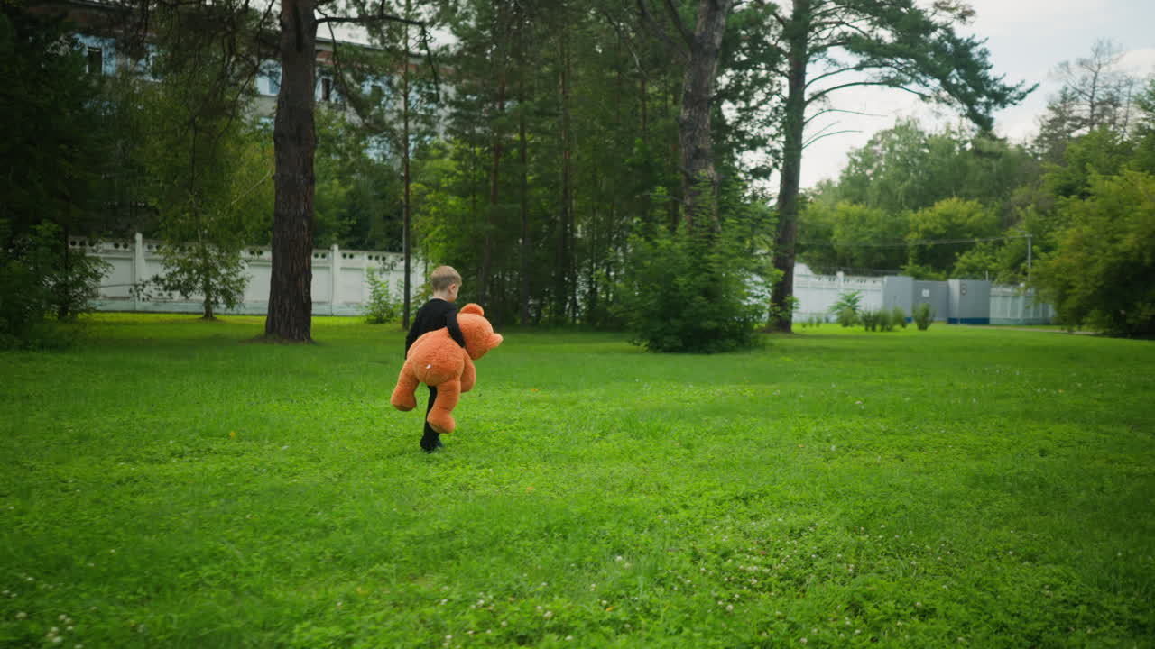 Rear view of young boy walking through green grassy area carrying large orange teddy bear, heading toward white fence with trees and official-style building in background under daylight sky