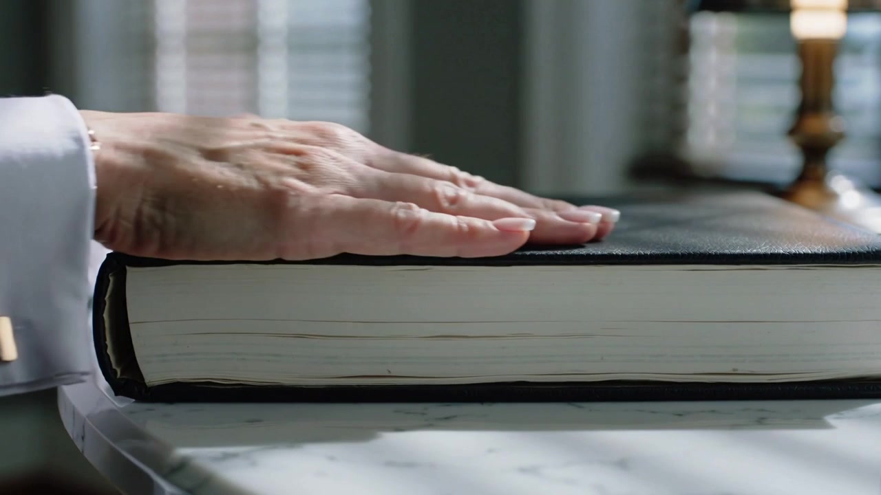 Sollevando le mani con i polsini, posa e liscia un libro nero su un ripiano in marmo vicino alle persiane per appiattirne la copertina.