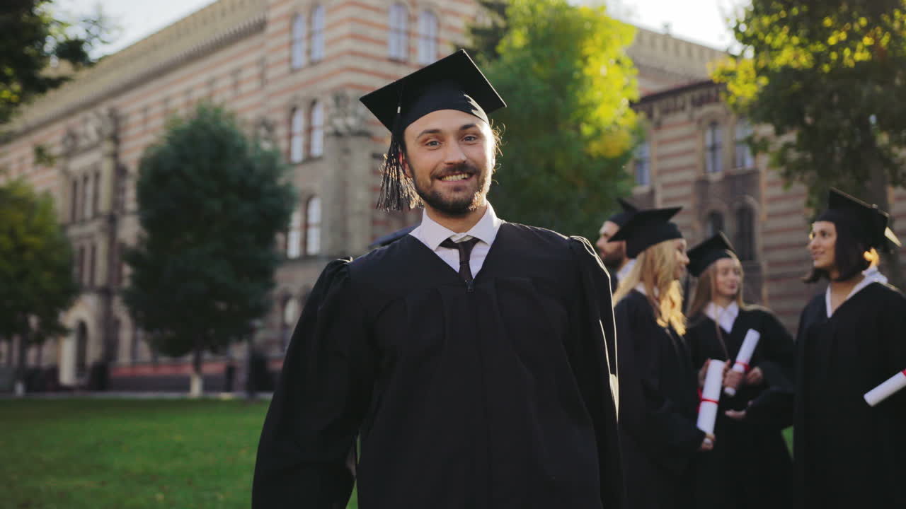 Portrait shot of the happy cheerful and funny man in traditional graduation gown and cap with diploma in hands having fun and grimacing to the camera