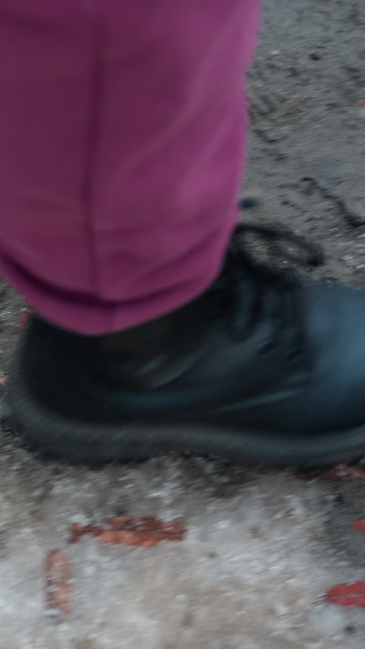 primer plano de la parte inferior de las piernas con pantalones marrones y botas negras caminando por un camino desgastado y helado cubierto de follaje esparcido y terreno accidentado y irregular, los pasos dejan huellas en el suelo congelado