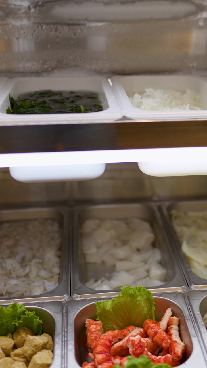 La telecamera scorre su vassoi di noodles crudi, verdure e ingredienti per hotpot sotto la luminosa illuminazione del buffet