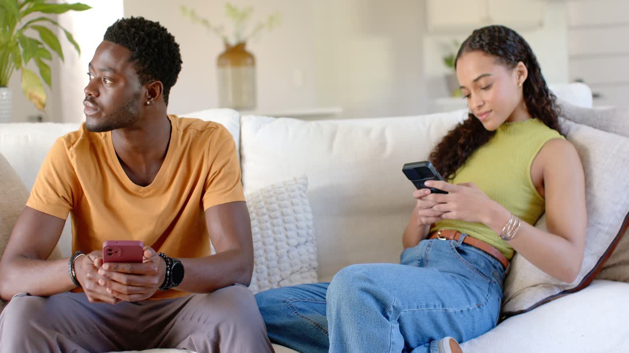 Casal afro-americano no sofá, a mulher enviando mensagens enquanto o homem, segurando um telefone vermelho, olha para a esquerda devido a um ruído.