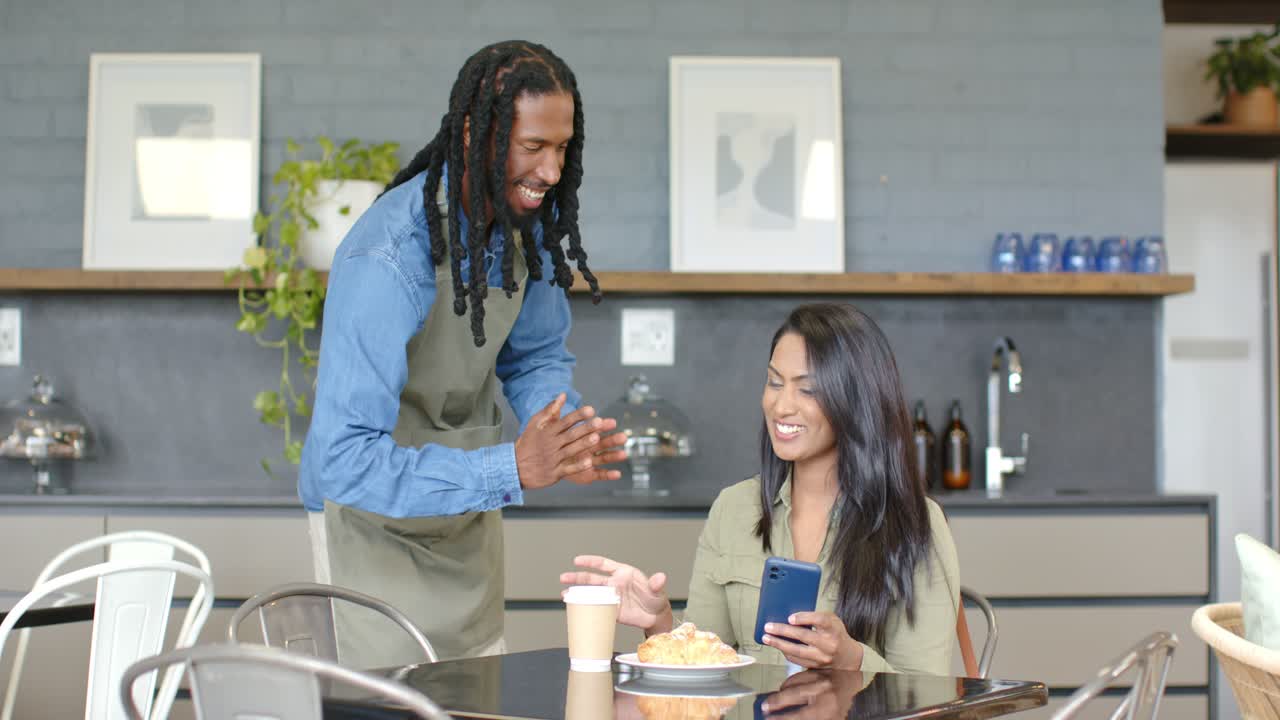 African American waiter serving coffee to Indian woman leaving her sipping and browsing smartphone