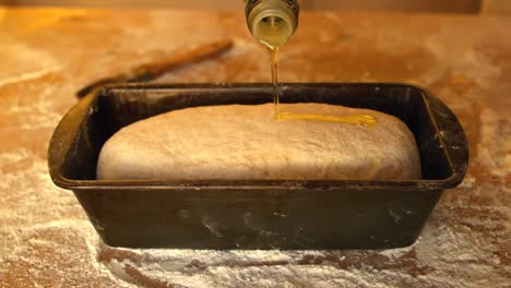 olive oil being poured over dough in loaf tin on a floury table