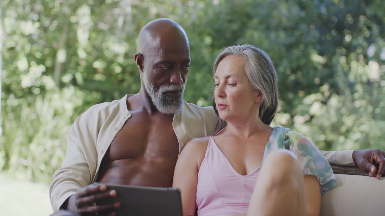 Couple sitting on patio sofa, tablet guiding man tapping as woman in pink top pointing, discussing
