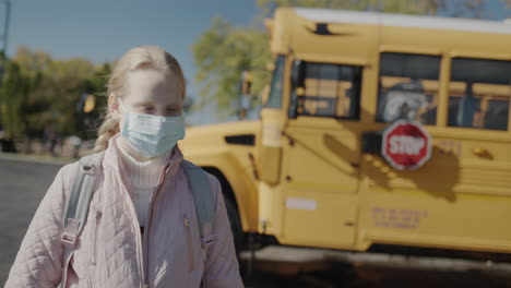 a schoolboy in a protective mask stands in front of a school bus. a stop sign is visible at the rear. protecting children from coronavirus concept