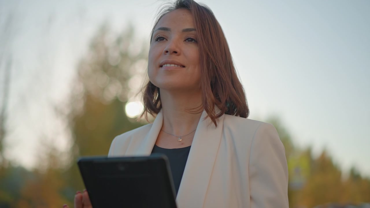 Jovem mulher confiante está ao ar livre sob a luz quente do sol, olhando para um tablet com um sorriso suave e uma leve inclinação da cabeça, mostrando foco calmo, envolvimento relaxado e um estilo de vida de trabalho moderno.