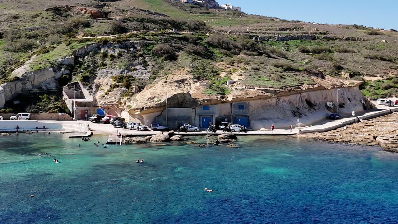 İç Deniz Dwejra Gozo'nun yüksek açıdan manzaralı görüntüsü; parlak güneş altında, kristal berraklığında turkuaz suyuyla, doğrudan engebeli kireçtaşı kayalıklarına oyulmuş geleneksel mavi kapılı kayıkhaneleri içeriyor