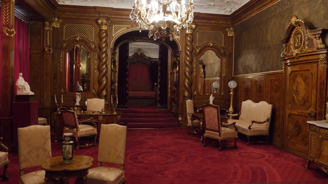 Ripresa panoramica di una sontuosa camera da letto reale all'interno del Castello di Peles, con colonne tortili in legno scuro, un letto a baldacchino, un tappeto rosso e un lampadario, a Sinaia.