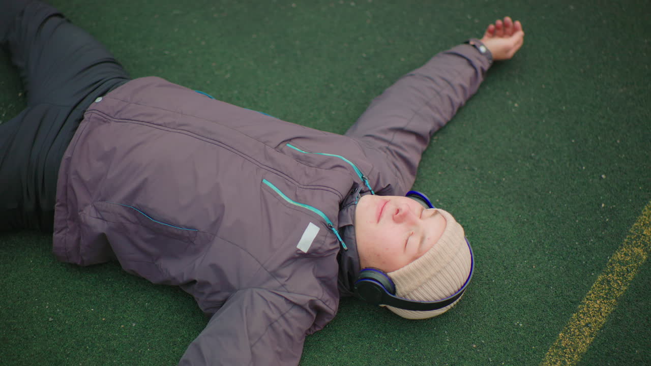 lying on turf with headphones, adult student takes reflective pause outdoors amid green field and yellow line, wearing winter jacket and beanie, arms outstretched, eyes closed in quiet mindfulness