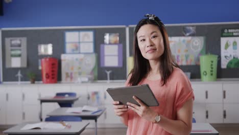 Portrait-of-happy-asian-female-teacher-with-tablet-in-elementary-school-classroom,-slow-motion