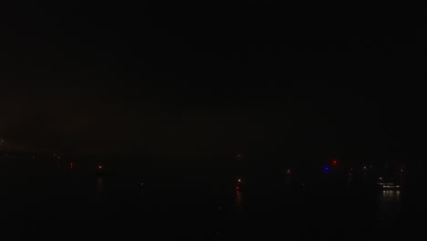 Fireworks-at-night,-lights-diffusing-in-cloudy-fog-over-ocean,-aerial