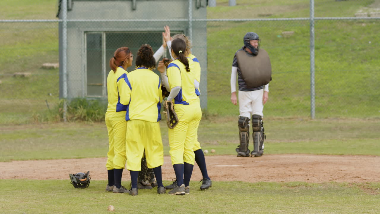 Multiracial female baseball players and male umpire huddling and celebrating on a pitch