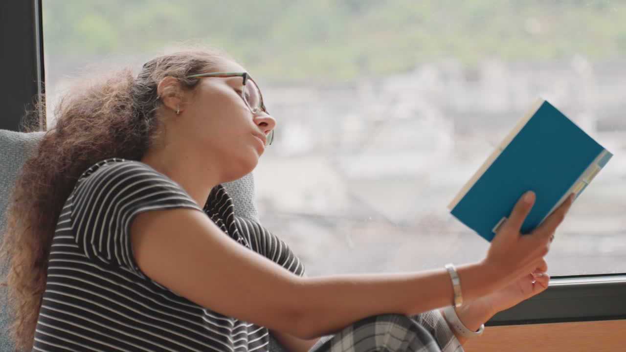 Nahaufnahme einer Frau, die ein blaues Buch auf einer Hotelsimse liest, mit nachdenklichem Blick und Brille, Armband-Detail und verschwommenem Stadtpanorama draußen, intimes Porträt, das einen ruhigen Rückzug und Nachdenklichkeit einfängt.