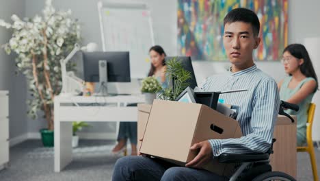 disabled sad men in shirt with asian korean beauty sits in wheelchair at desk in the company on knees holding cardboard box with packed things, gets fired from job unable to fulfill his duties