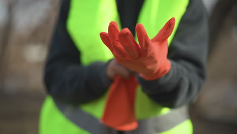 worker in dark sweater and high visibility vest putting on bright orange protective gloves outdoors, preparing for safe work in industrial or construction environment, focusing on hand safety gear