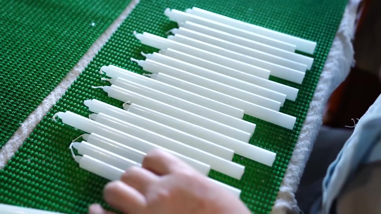 Worker arranging rows of tapered candles on a green mesh mat beside branded boxes at a factory