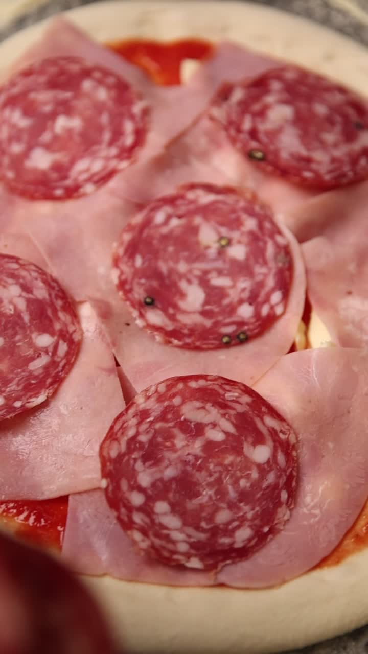 Mano che posiziona fette di salame su una pizza con prosciutto e salsa