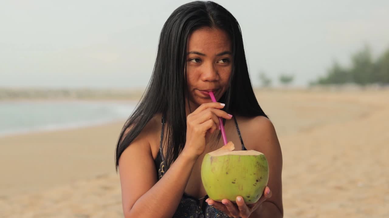 una joven asiática está bebiendo un refrescante refresco de coco natural en la playa