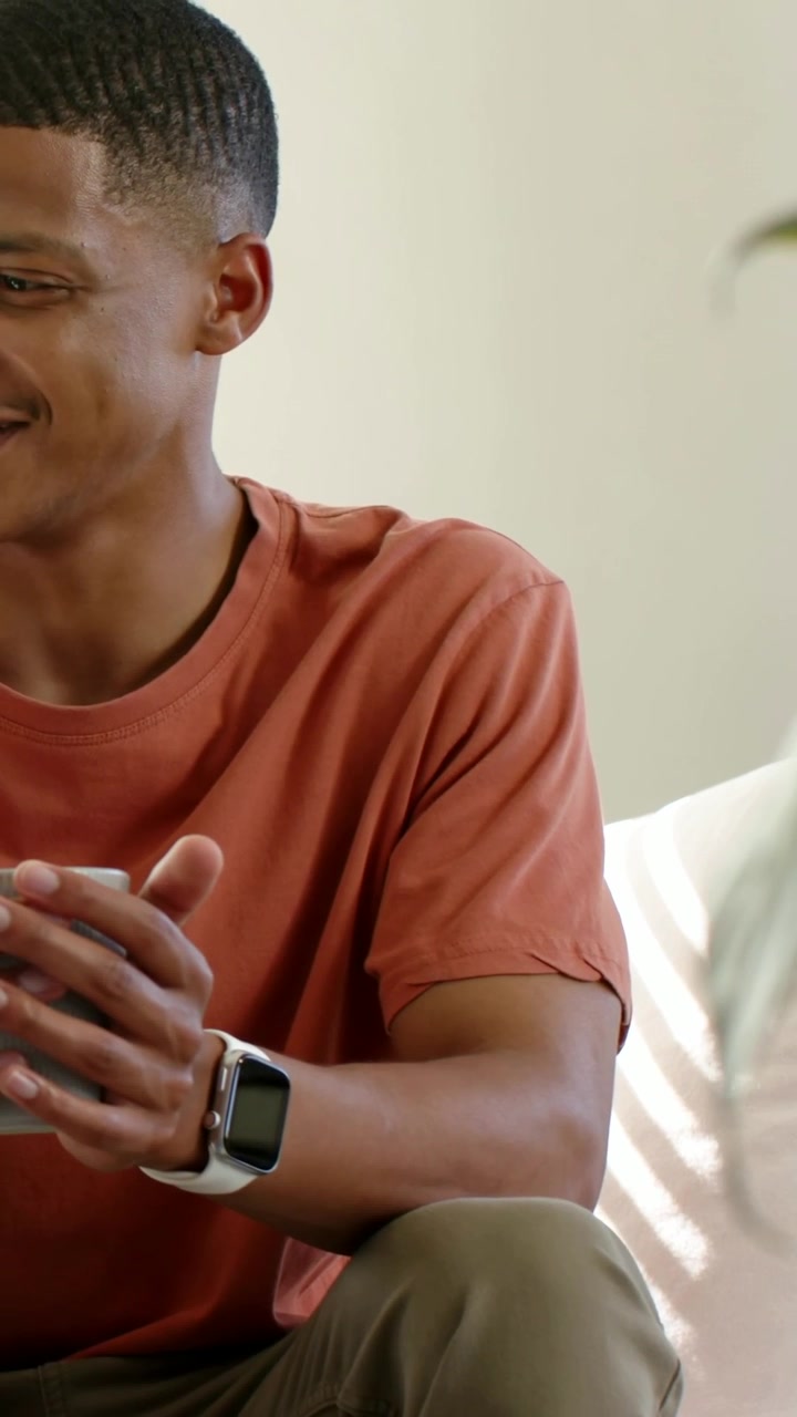 Vertical video: Smiling man in rust tee shifting hearing offscreen voice on sofa, with smartwatch