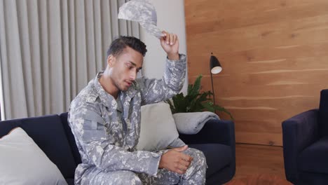 soldado masculino biracial serio en uniforme sentado en el sofá en casa haciendo una videollamada, cámara lenta