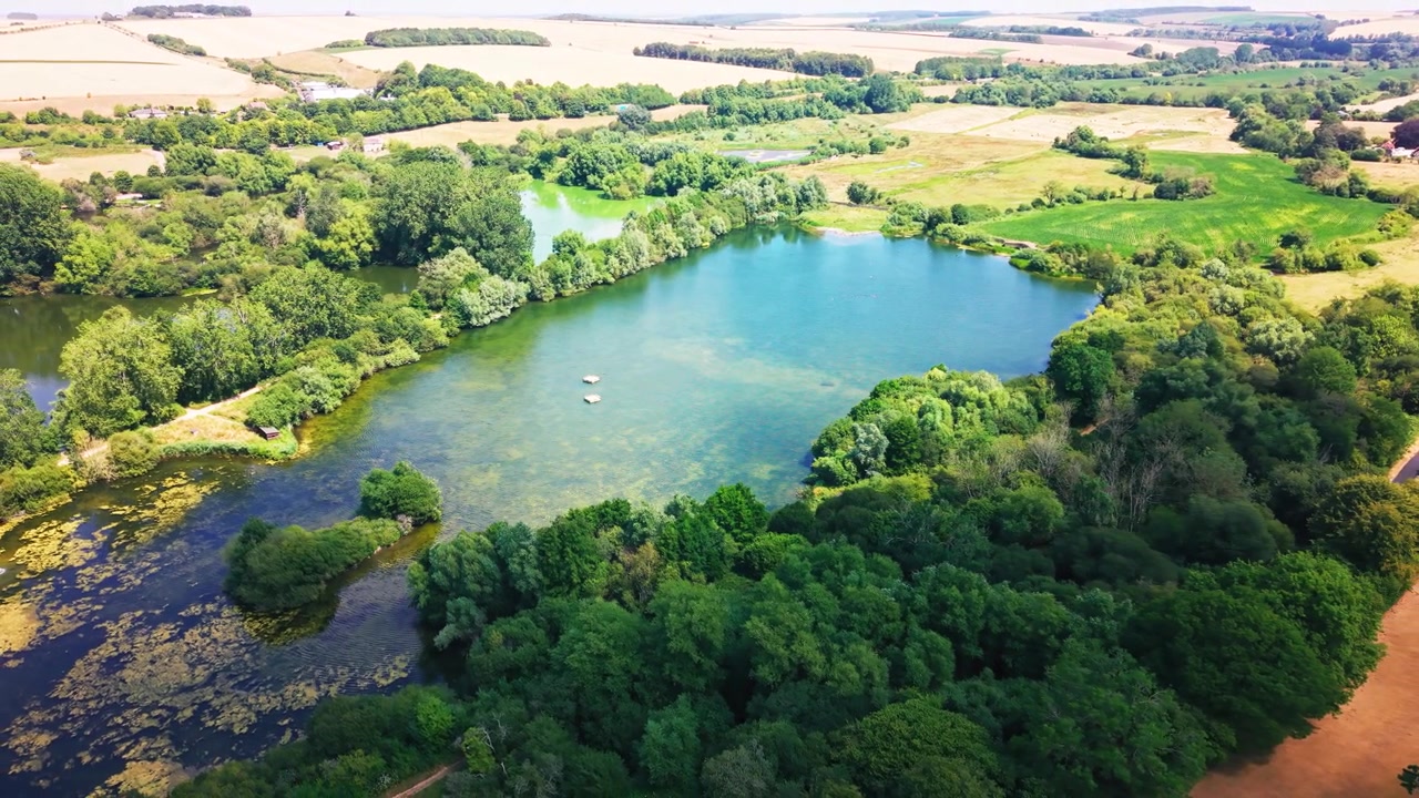Panoramische dronebeelden van een helder zoetwatermeer, omringd door dicht bos, moerasvegetatie en open landschap, bij natuurreservaat Langford Lakes in Wiltshire, Engeland.