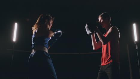 Schöne-Brünette-Boxerin-Trainiert-Mit-Einem-Trainer-In-Einem-Dunklen-Raum-Schläge-Auf-Die-Pfoten