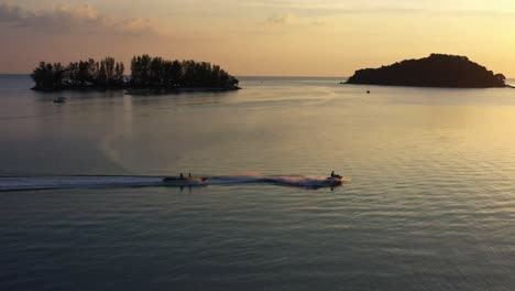 Atemberaubende-Kamerafahrt-Aus-Der-Luft,-Die-Einen-Jetski-Einfängt,-Der-Ein-Bananenboot-Hinter-Sich-Herzieht,-Zur-Goldenen-Stunde-Des-Sonnenuntergangs-über-Das-Wasser-Kreuzt,-Unterhaltsame-Wassersportaktivitäten-Auf-Der-Insel-Langkawi,-Malaysia