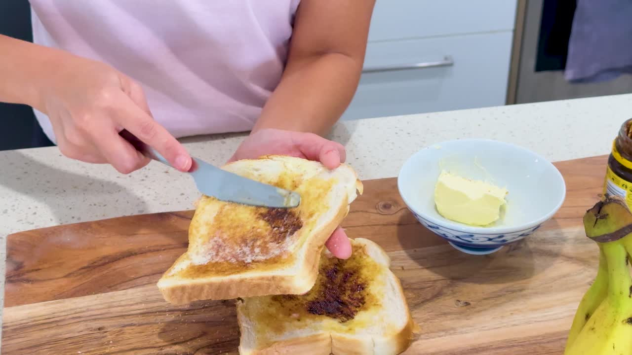 A person spreads butter onto slices of toasted bread with dark yeast spread on a wooden board in a brightly lit modern kitchen