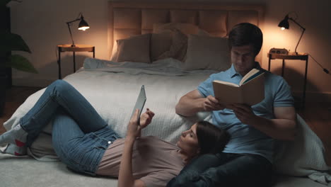 young couple sitting in bedroom, woman and man reading a tablet and a book and relaxing, sharing moments