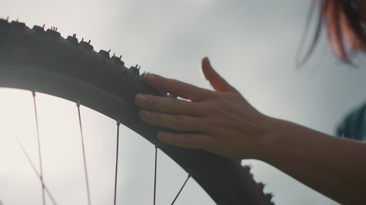 woman touching bike tire against sky, soft backlight closeup of hand on muddy mountain tire, slow cinematic movement, tactile exploration, reflective mood, outdoor trail, late afternoon light, quiet