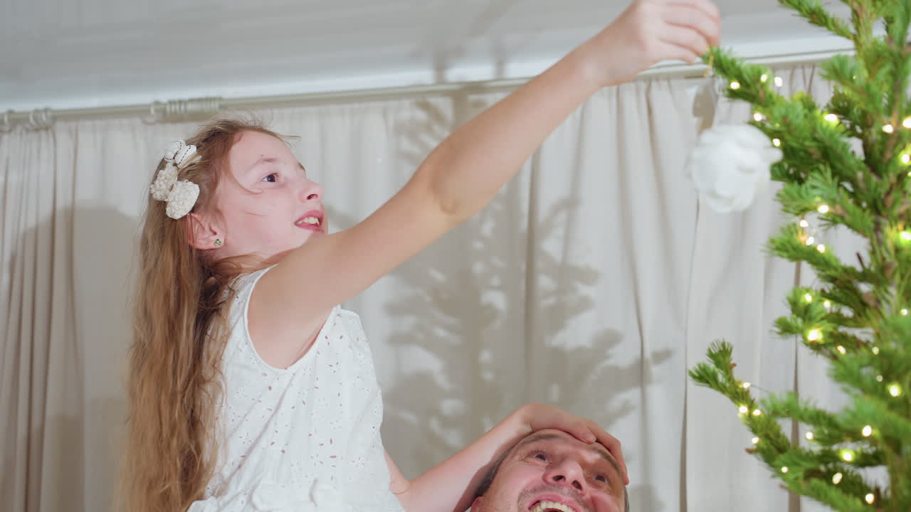 Close up of young daughter in white gown holding dads head lovingly while reaching out to place beautiful white ornament on decorated christmas tree together inside cozy home during festive holiday