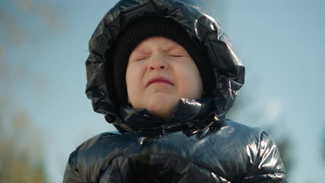 a close-up of a young child wearing a shiny black jacket, with a hood and a beanie, looking slightly frustrated and squinting as he open his eyes slowly