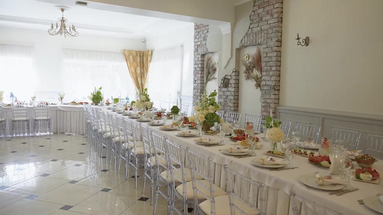 Mesa de banquete bellamente dispuesta en un salón de eventos moderno y soleado, con elegantes centros de mesa florales, sillas de cristal y un ambiente acogedor y festivo sin personas presentes