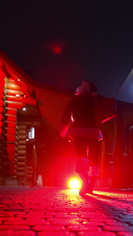 Santa Claus with bag full of presents. Santa Clause walking to the decorated houses with lights and carrying Christmas gifts. Red lighting. Back view. Vertical video