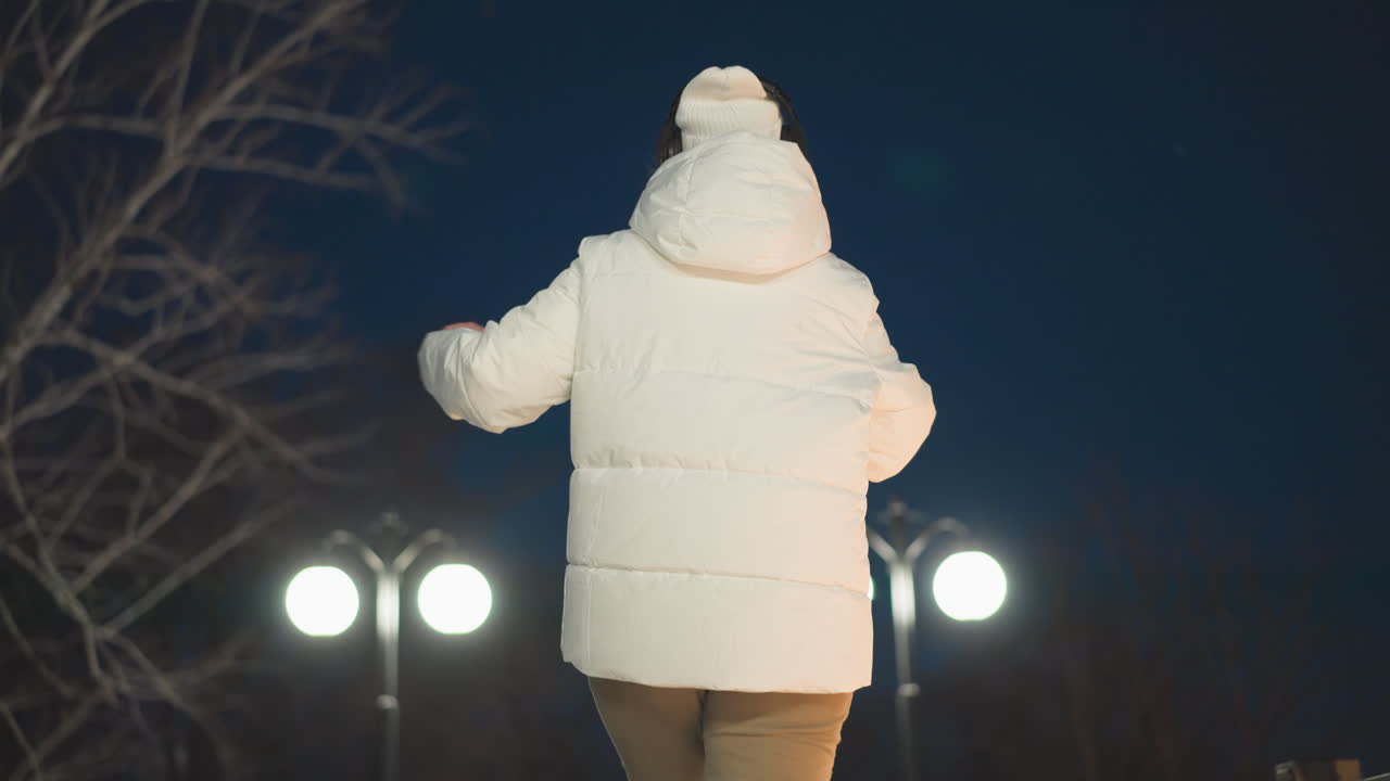 Back view of woman in white puffer coat and headphones swaying to music on snow covered walkway under bright lampposts at night expressive movement in winter park scene with peaceful ambient mood
