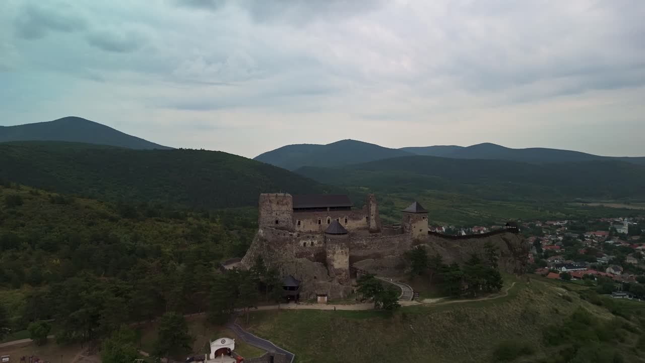 Backward tracking aerial view of Boldogkő Castle in Boldogkőváralja, Hungary, perched on a hilltop amid lush green hills and forests under cloudy skies, showcasing medieval architecture