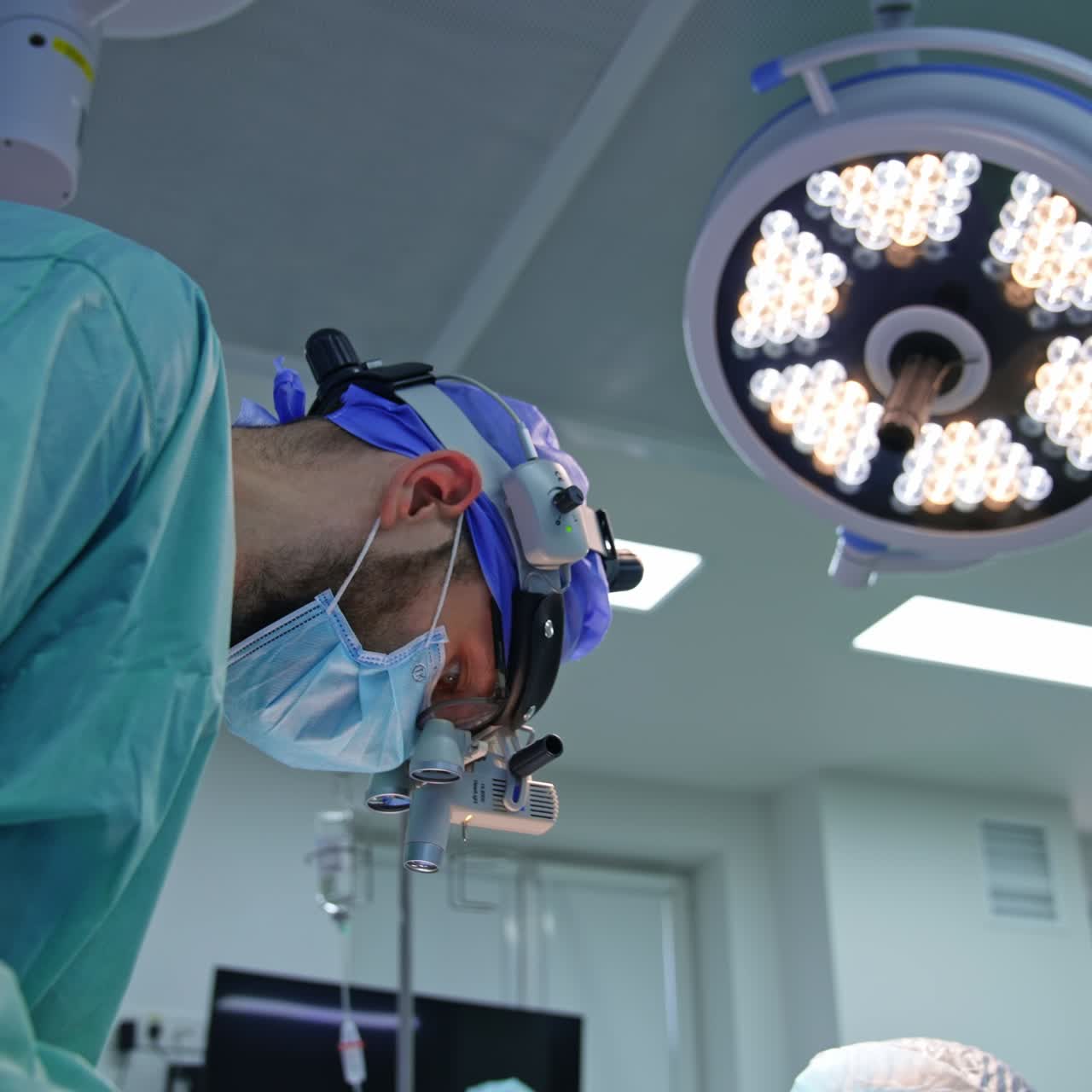 Young mature male doctor in device glasses on his head bent over the patient. Surgeon working under the big lamp in modern surgery room