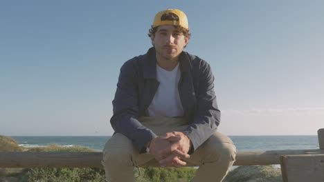 hombre guapo con gorra sentado en el paseo marítimo mirando a la cámara