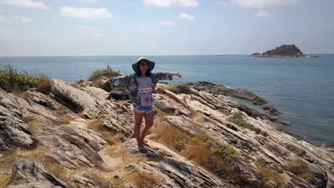 aerial: a drone rotates around a young thai woman that is standing at a very high point on some rocks and stones and rises her arms in the air smiling and closing her eyes