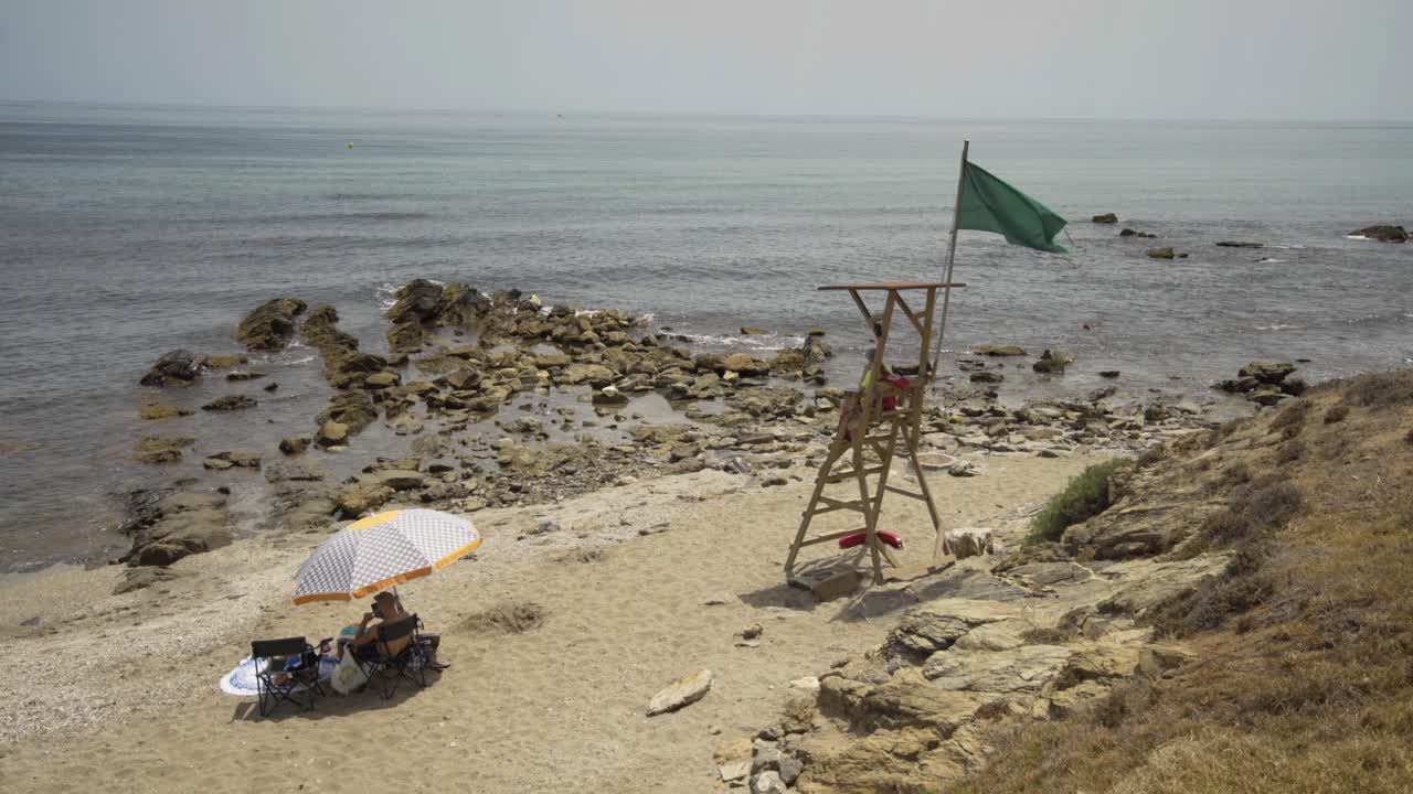 Life guard sits on duty on the beach near Cala de Mijas on the Costa Del Sol in Southern Spain while a holiday makers sits beneath parasol.
