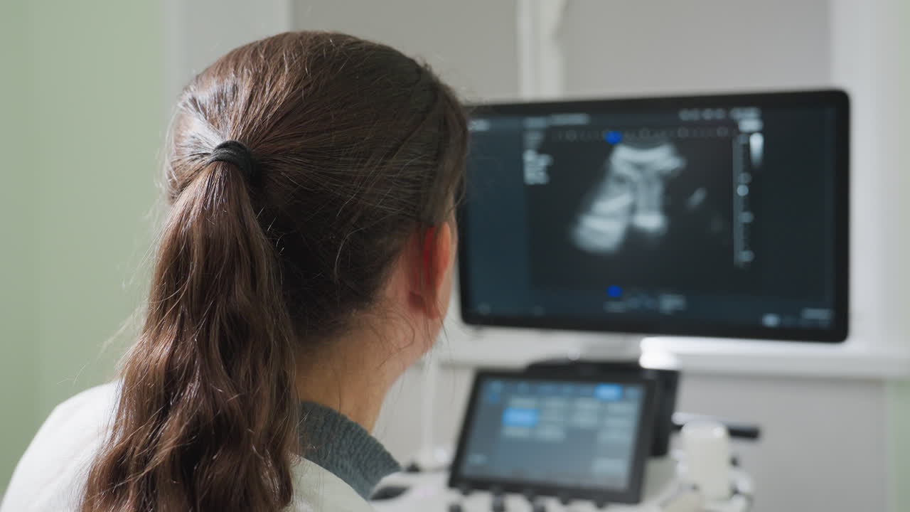 Lab researcher with tied hair observes images on monitor of MRI machine in clinical setting, focusing on detailed black and white scan display for diagnostic analysis and data interpretation