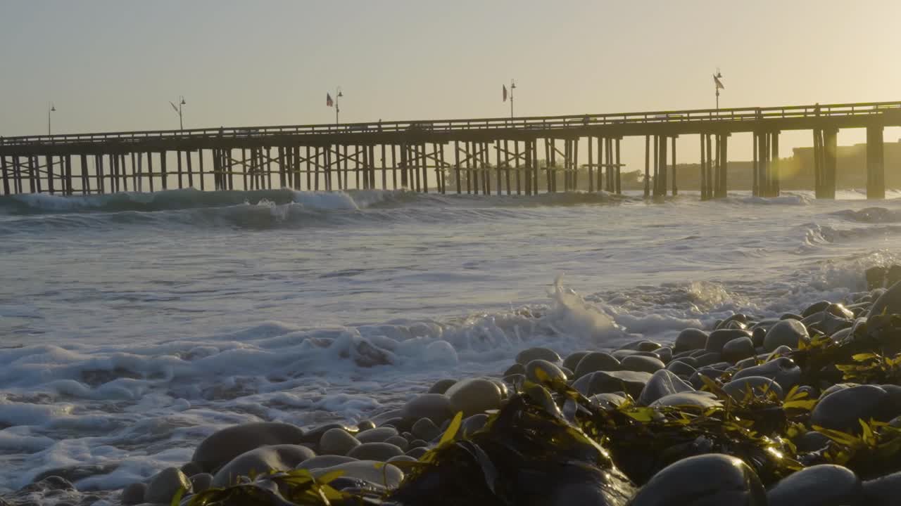 Waves roll onto a rocky shoreline covered in seaweed as the sun sets behind a wooden pier stretching into the ocean