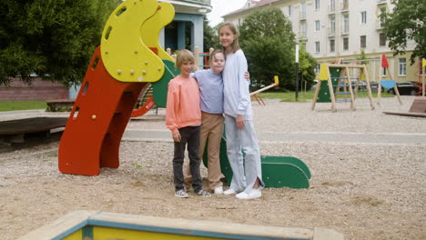 little girl with down syndrome hugging another girl and little boy in the park on a windy day. they are looking at camera