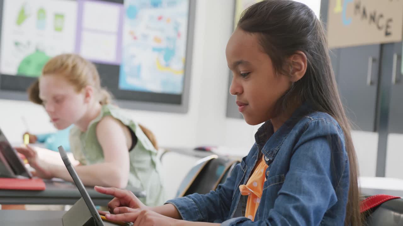 Video of smiling biracial schoolgirl sitting at desk using tablet in diverse class, copy space